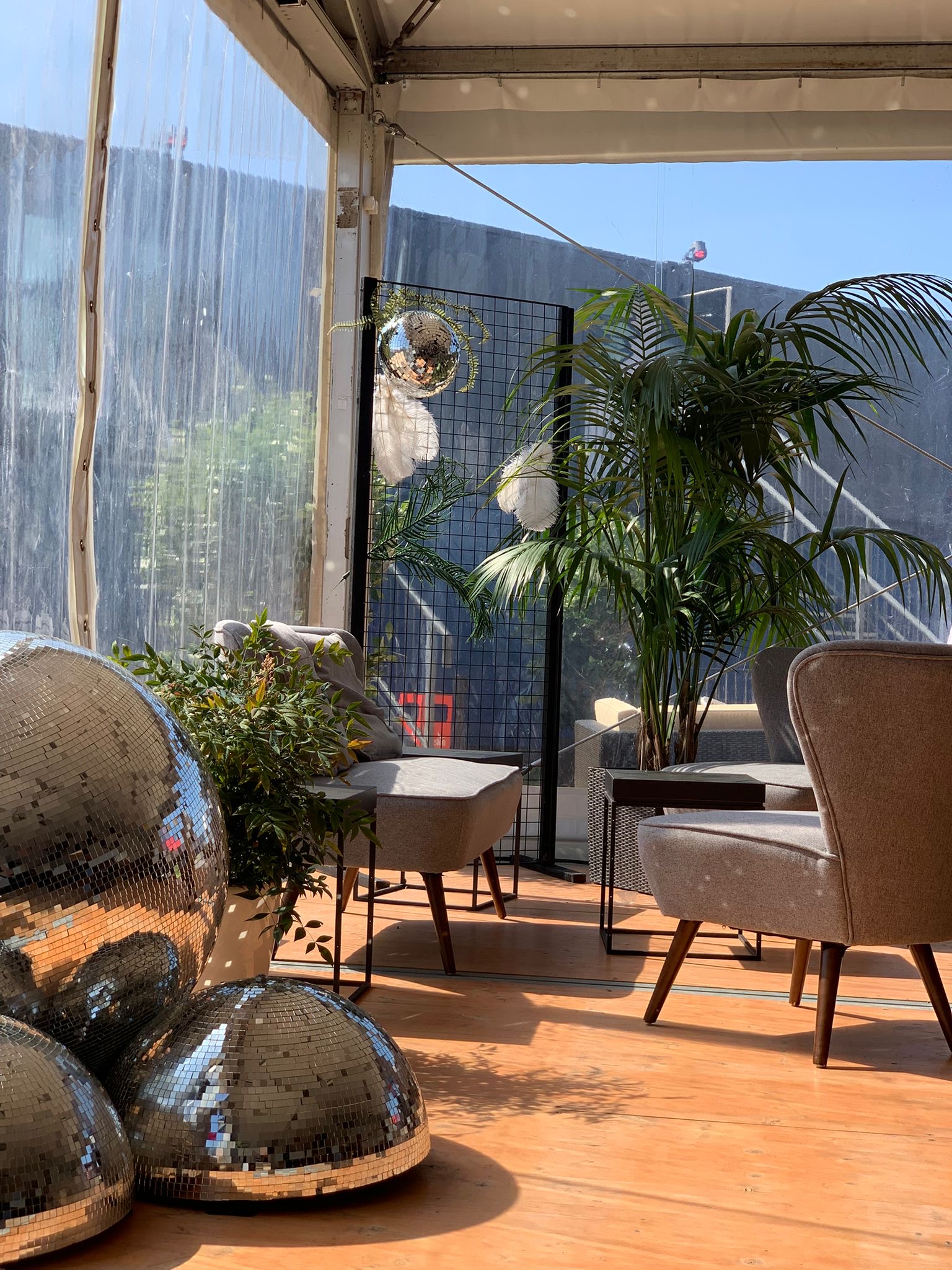 Moderne Event-Lounge mit grauen Sessel-Stühlen, Palmen, schwarzen Gitter-Raumteilern und großen Diskokugeln auf Dielenboden von Event Rent.