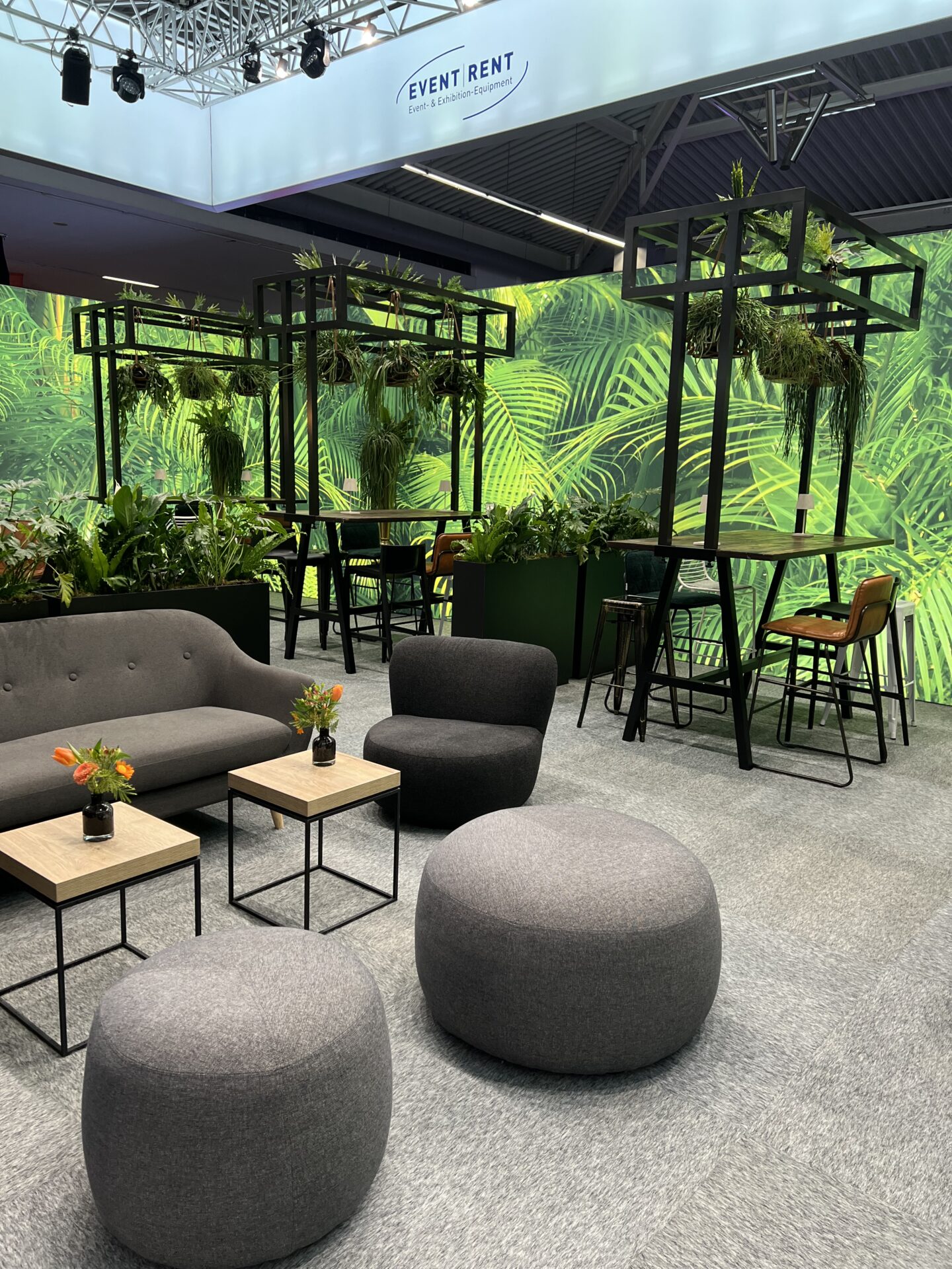 Messestand von Event Rent mit grauer Loungeecke, Stehtischen und Urban Jungle Wanddeko.