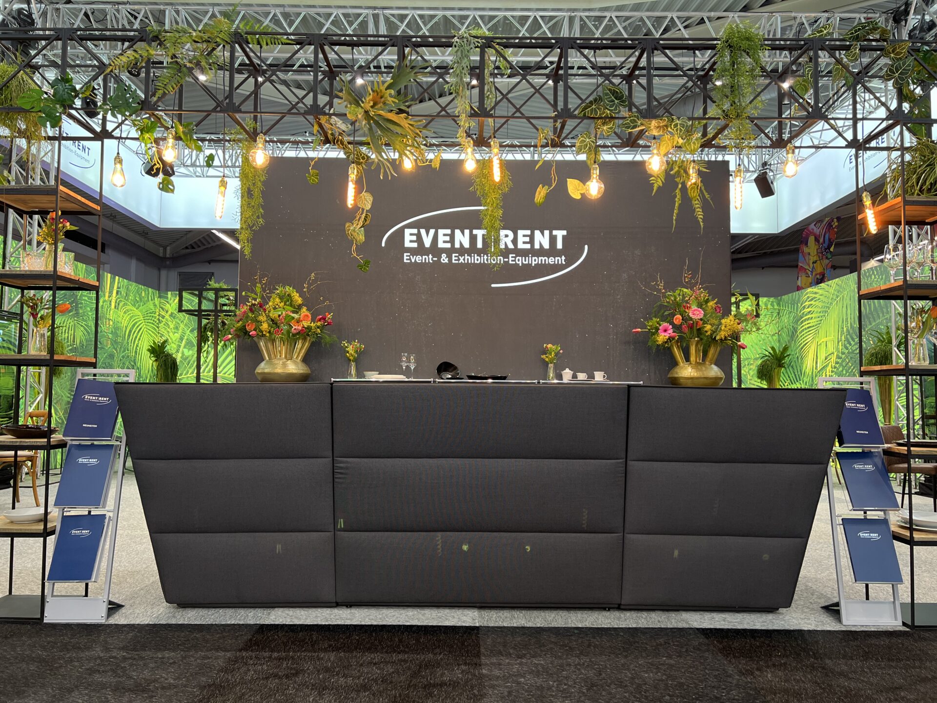 Eigener Messestand von Event Rent mit Empfangstresen, goldenen Blumenvasen und Urban Jungle Deko.