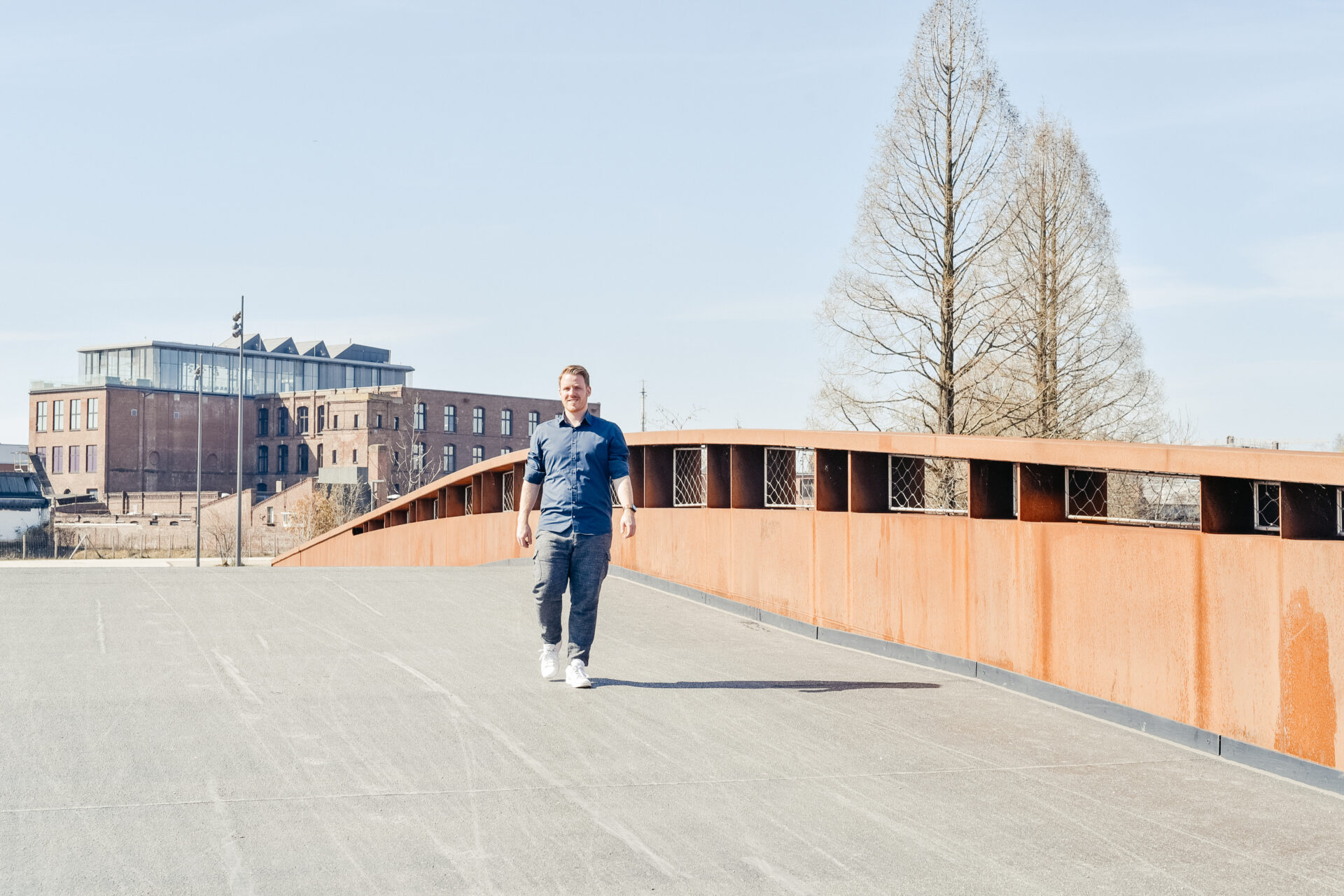 Jens Bungert von Event Rent geht in einem blauen Hemd und dunkler Hose über eine Fußgängerbrücke vor moderner Industriearchitektur.
