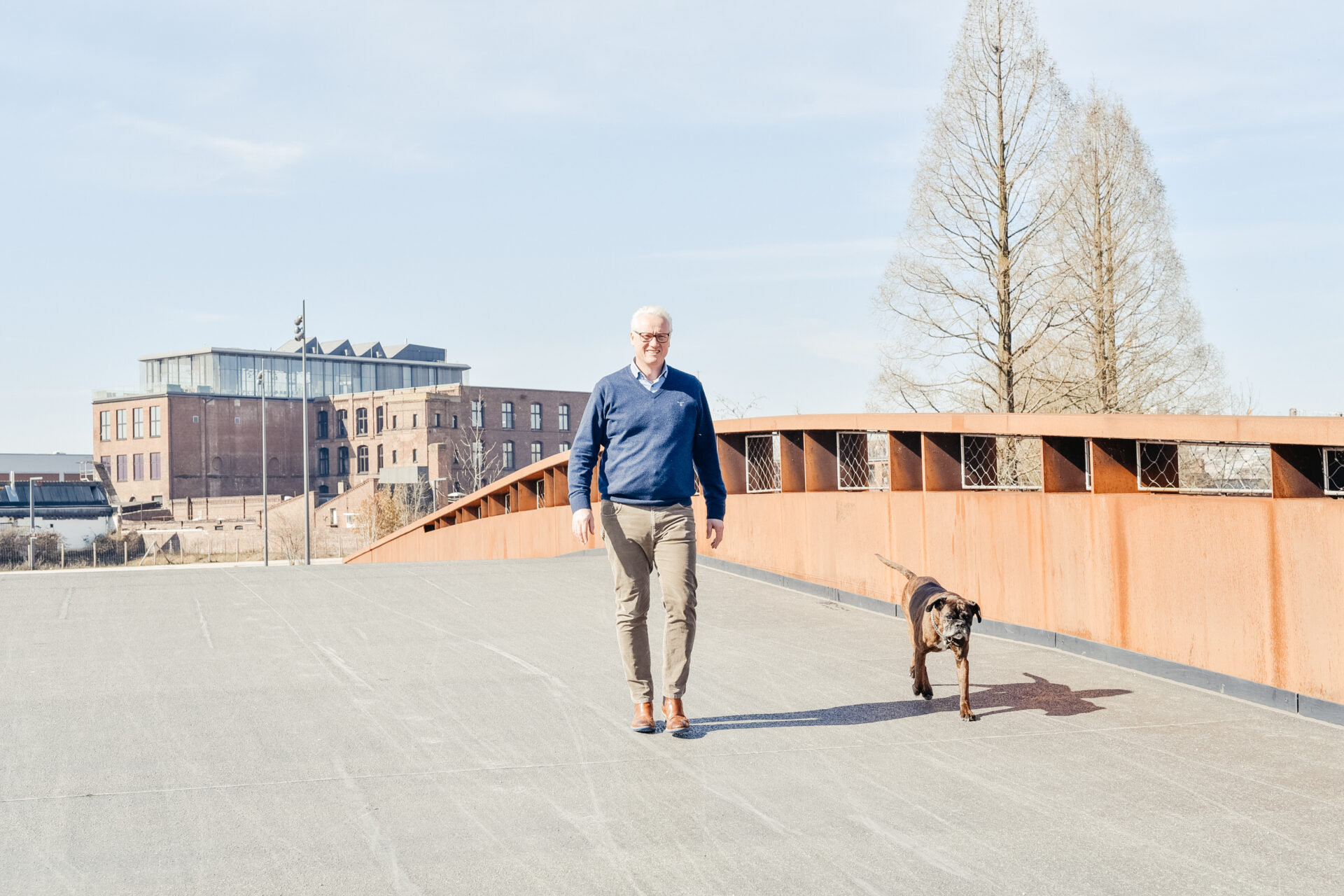 Jürgen Bungert von Event Rent geht zusammen mit Hund Flocke über eine Fußgängerbrücke vor moderner Industriearchitektur.