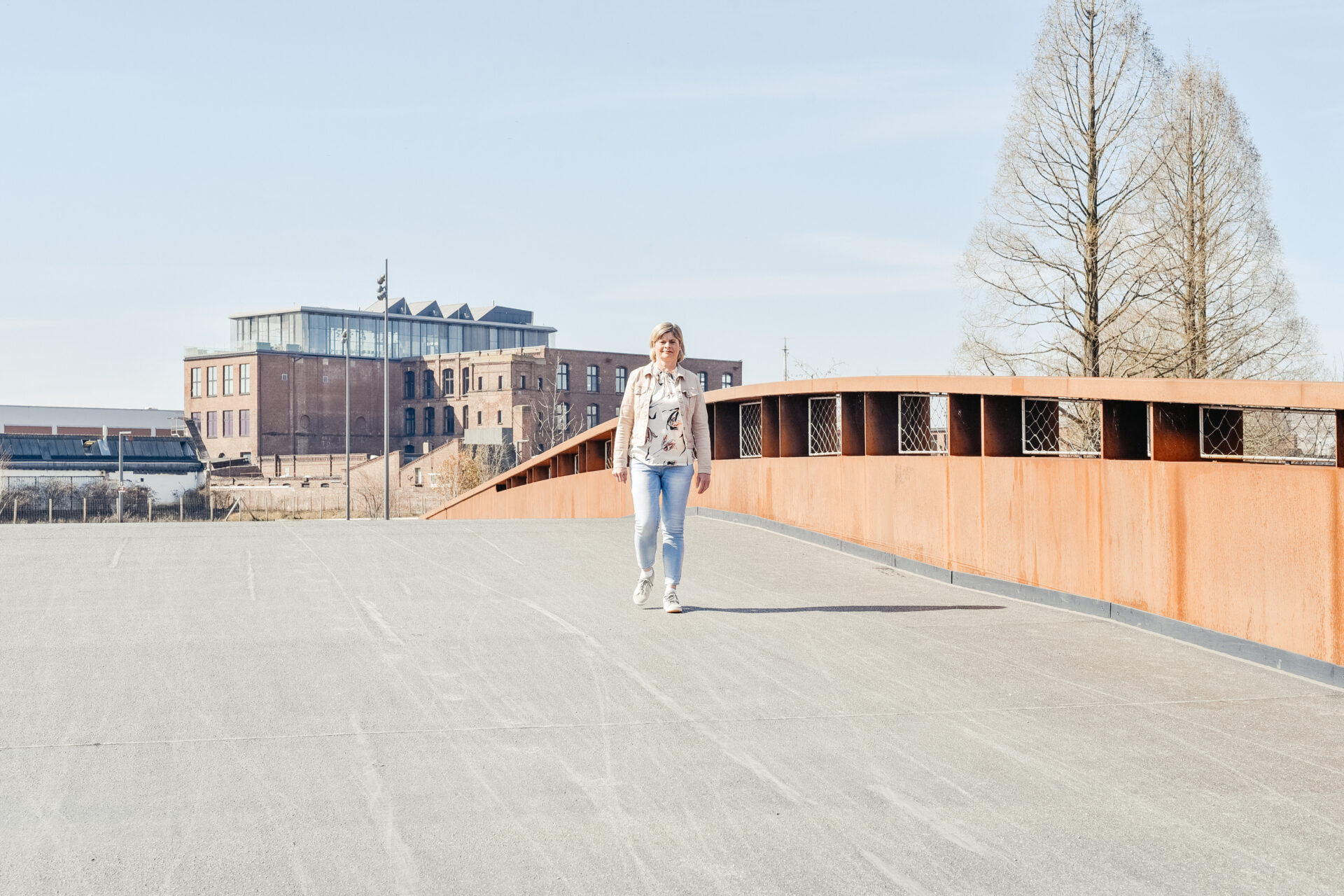 Renate Burian von Event Rent geht in hellem Outfit über eine Fußgängerbrücke vor moderner Industriearchitektur.