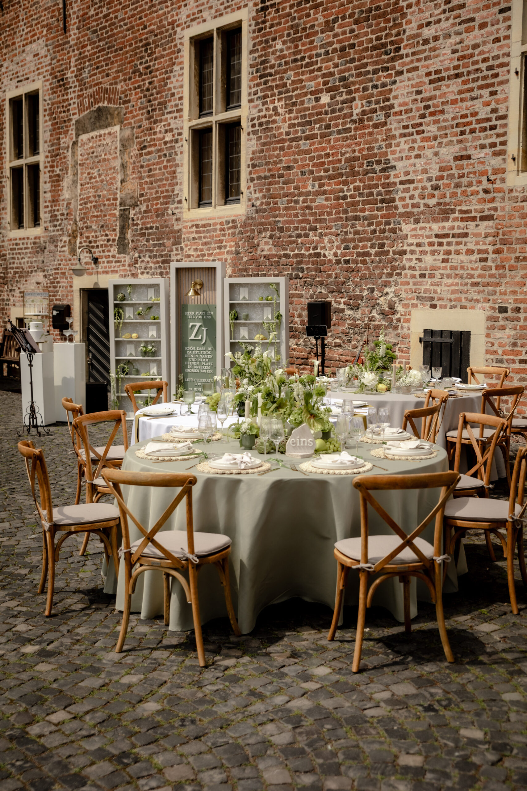Runder Banketttisch von Event Rent mit salbeigrüner Tischdecke, Crossback-Holzstühlen und üppiger Blumendekoration auf einem historischen Kopfsteinpflaster-Innenhof.