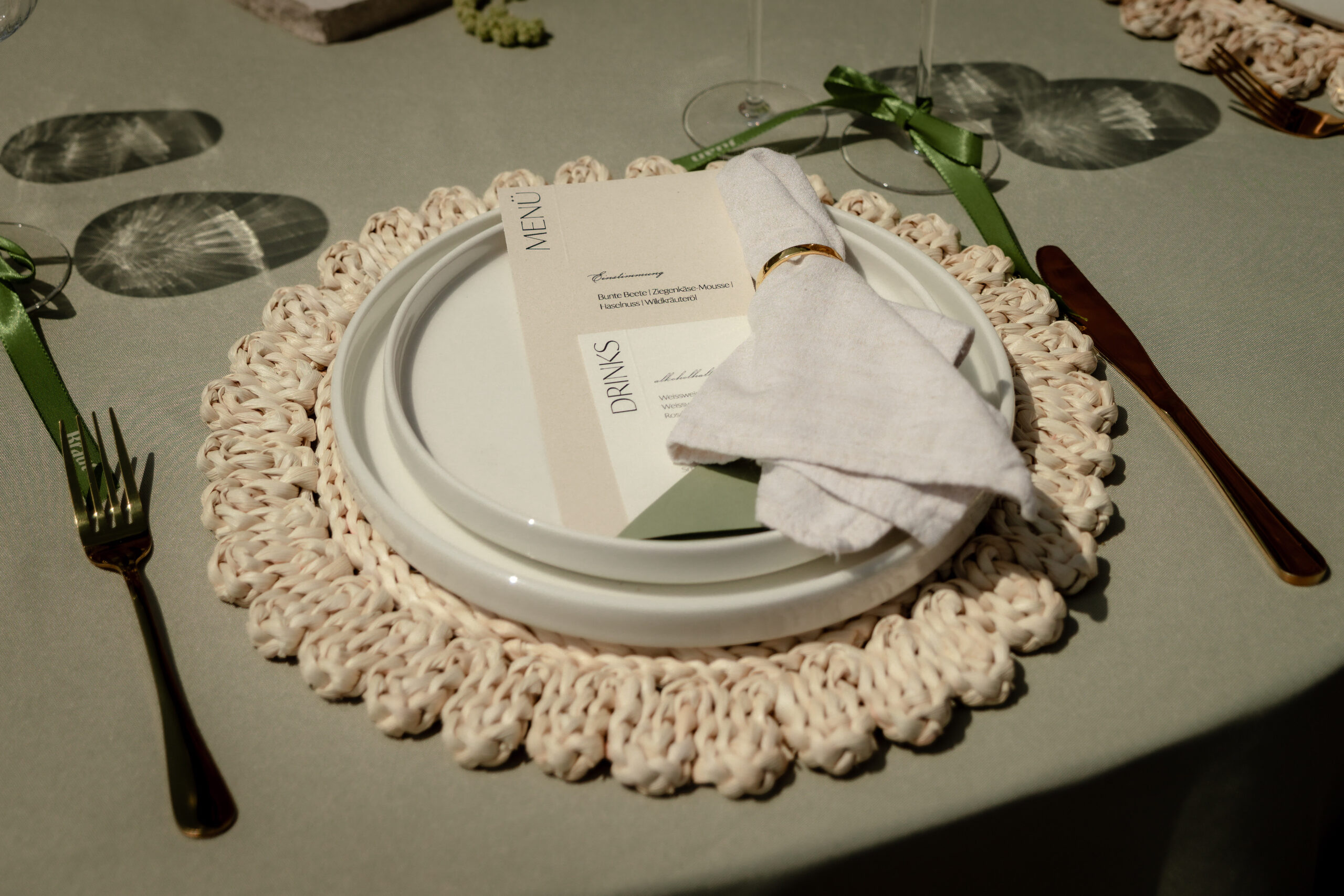 Elegantes Tischgedeck auf salbeigrüner Tischdecke von Event Rent mit Makramee-Platzset, weißen Tellern, Stoffserviette, Menükarte und goldenem Besteck.